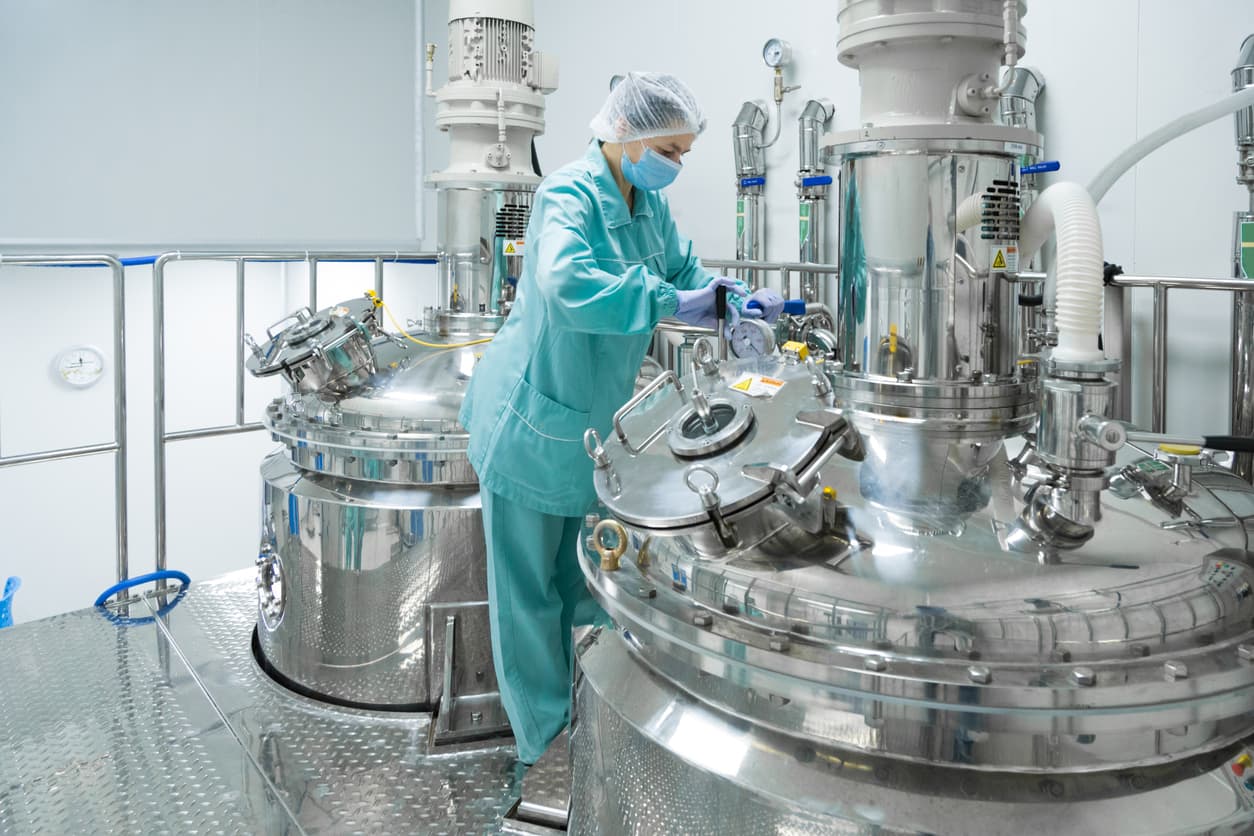 Técnica en laboratorio farmacéutico ajustando equipos en una planta de procesamiento con tuberías de acero inoxidable, destacando la precisión y la higiene en la manufactura.